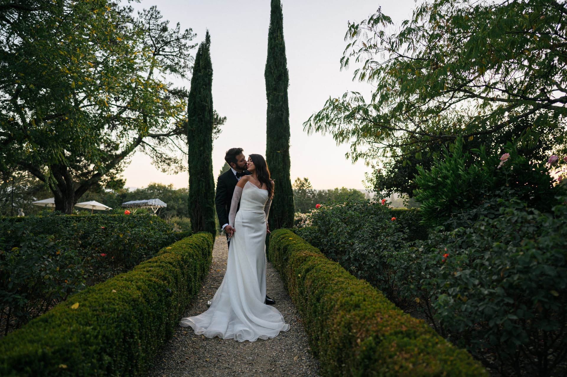 Bride and groom embracing on a hedge-lined garden path at sunset