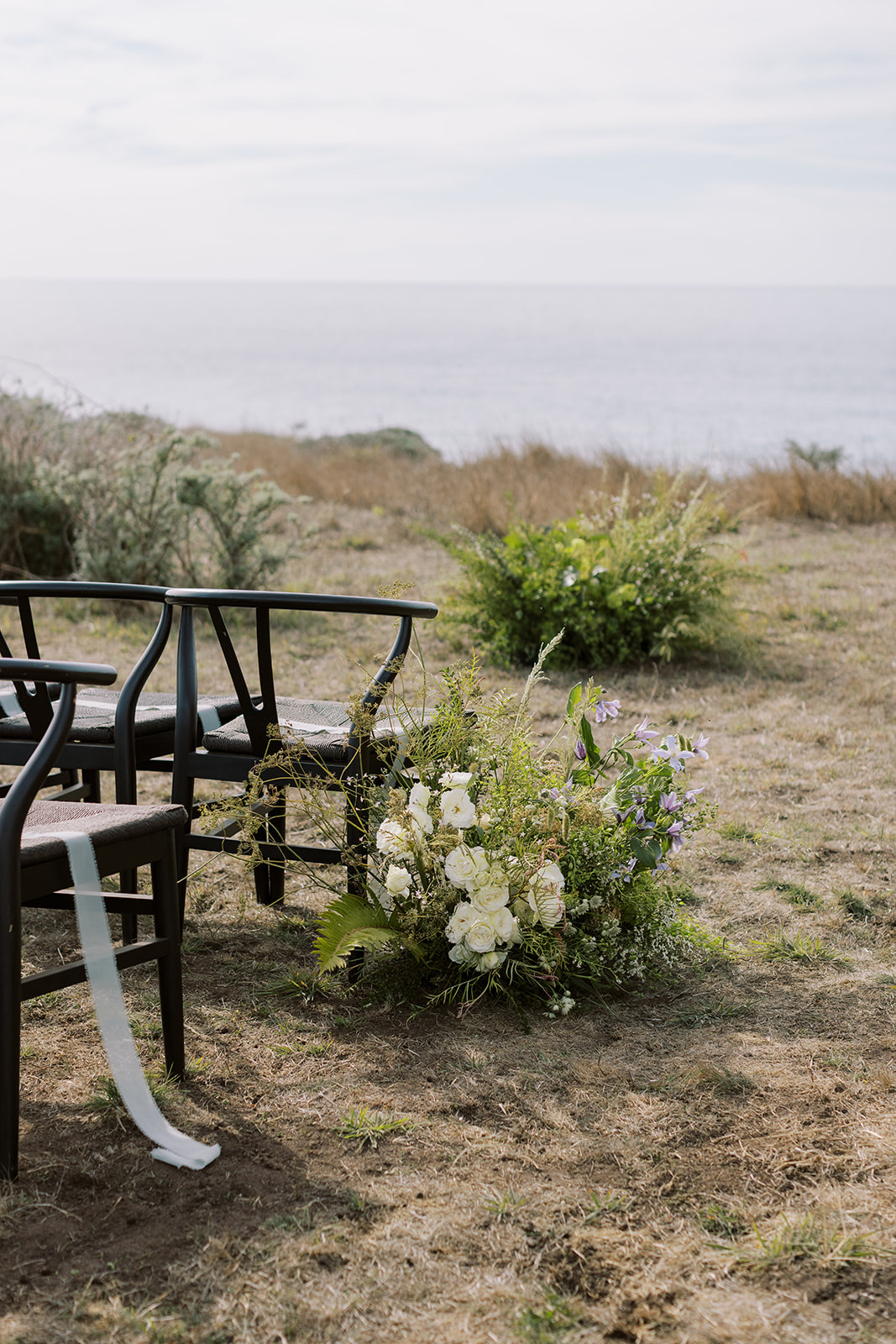 Wild white and lavender ceremony florals beside black wishbone chairs on a coastal bluff